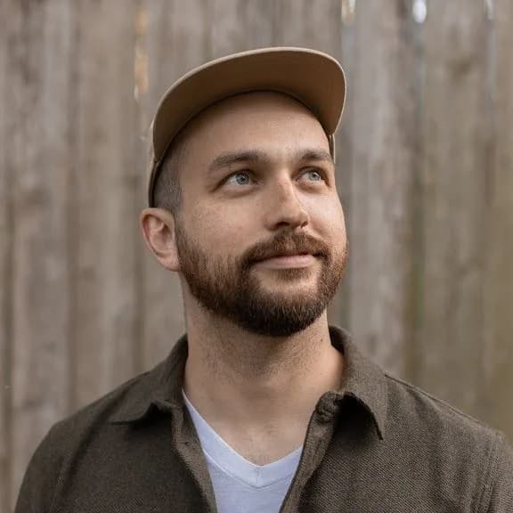 a man with a beard and a hat is standing in front of a wooden fence .
