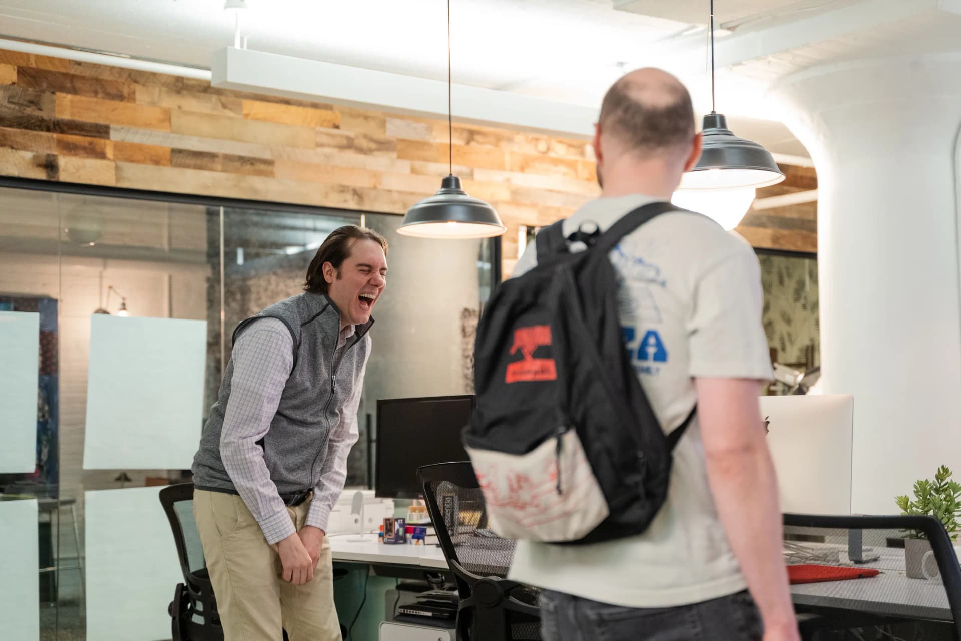 a man with a backpack is laughing with another man in an office .